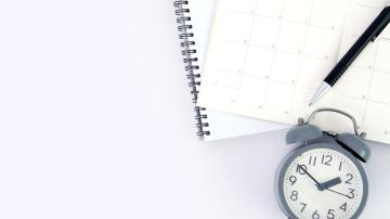 Pen on spiral notebook and calendar with alarm clock below it