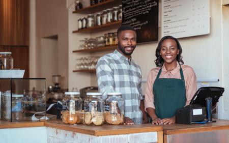 Portrait of two smiling young African entrepreneurs standing welcomingly together behind the counter of their trendy cafe